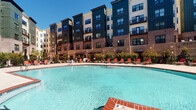 Located at 550 Pitt Greene Connector in Greenville, North Carolina, this outdoor amenity area serves as a central gathering space within a modern, multi-story residential community. The focal point is a generously sized resort-style swimming pool with clear blue water, framed by a broad concrete sundeck that provides ample room for lounging and socializing. Red lounge chairs and umbrellas are arranged throughout the deck, offering shaded and sun-lit seating options for residents. Brick-accented pool coping adds a classic touch to the contemporary design, complementing the mixed-material exterior of the surrounding building, which features dark panel siding, brick facades, and large windows with private balconies on upper floors. Mature landscaping, including ornamental trees and manicured shrubs, lines the perimeter of the courtyard, softening the architectural edges and adding greenery to the space. Wrought iron fencing defines the boundaries of the pool area while maintaining open sightlines. The courtyard orientation allows for abundant natural sunlight throughout the day, enhancing the inviting, open-air atmosphere. A dedicated patio area with additional seating under shade umbrellas provides a quieter retreat within the amenity space, making it well-suited for both relaxation and social gatherings.