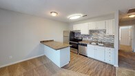 Located at 2000 Lions Gate Dr in Augusta, Georgia, this single-story residence offers a practical and well-appointed layout designed for comfortable everyday living. The open-concept kitchen serves as a central feature, equipped with stainless steel appliances including a refrigerator, electric range, and dishwasher. White cabinetry provides ample storage, while dark granite-style countertops and a mosaic tile backsplash add visual interest. A peninsula counter extends into the adjacent living area, creating a natural transition between spaces and offering additional workspace or casual seating. The living area is generously sized, finished with neutral carpet, and receives natural light through a set of double doors with flanking windows. Hardwood-style flooring runs through the kitchen and bathroom areas, providing a cohesive contrast to the carpeted rooms. The bedroom is a well-proportioned space with soft carpeting and neutral walls, accompanied by a spacious walk-in closet outfitted with wire shelving and hanging rods on multiple walls. The bathroom includes a single-vanity with white cabinetry and granite-style countertop, a tub-shower combination, and a pendant light fixture. Throughout, textured ceilings and neutral gray walls contribute to a clean, move-in ready aesthetic.