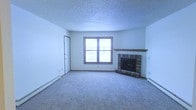 This single-floor space presents a clean and functional layout designed for comfortable everyday living. The main living area is a generous, open room finished with wall-to-wall carpeting that adds warmth underfoot. A standout feature of the room is the brick fireplace situated in the corner, complete with a wood mantel, serving as a natural focal point and providing a cozy atmosphere during cooler months. A centered window allows natural light to fill the space, brightening the neutral white walls and textured popcorn ceiling. Baseboard heating units are present along the walls, ensuring efficient temperature control throughout the space. A interior door provides access to additional areas of the floor plan, contributing to a practical and connected layout. The overall ambiance is understated and neutral, offering a blank canvas that lends itself well to a variety of interior styles. The combination of classic architectural details — including the brick fireplace surround, carpeted floors, and textured ceiling — gives the space a traditional residential character that is both welcoming and easy to personalize.
