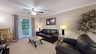 This single-floor residence offers a comfortable and functional layout across several well-defined living areas. The living room serves as the central gathering space, featuring plush carpeting, crown molding, and a ceiling fan overhead. A sliding glass door floods the room with natural light and provides direct access to an outdoor patio with views of surrounding greenery. The space comfortably accommodates a full seating arrangement alongside a glass-top coffee table and a dedicated media area with built-in mirror accent and TV stand.

The kitchen is efficiently designed with ample cabinetry, light countertops, tile flooring, and a full suite of black appliances including a range, over-the-range microwave, dishwasher, and refrigerator. Adjacent to the kitchen is a cozy dining nook with tile flooring and a door leading to the patio, ideal for casual meals.

The primary bedroom is generously sized with soft carpeting, a ceiling fan, and a large window allowing natural light. It features rich wood furniture including a sleigh bed and coordinating dresser with mirror. The en-suite bathroom offers an extended vanity with a large mirror, tile flooring, and plantation shutters. A second bathroom includes a tub-shower combination with chrome fixtures and tile flooring.