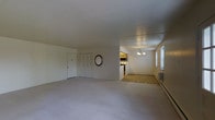This single-floor residence offers a straightforward and functional layout designed for comfortable everyday living. The main living area is generously proportioned, featuring soft, neutral-toned carpeting that runs throughout the space, contributing to a calm and understated ambiance. A closet with bifold doors provides convenient built-in storage near the entry, keeping the living area clean and uncluttered.

An open passageway connects the living room to the adjoining dining area, which features hardwood-style flooring and benefits from natural light streaming in through a set of windows along the wall. An elegant chandelier adds a touch of warmth and character to the dining space, making it a welcoming area for meals and gatherings.

The kitchen is positioned just off the dining area and is equipped with overhead cabinetry and appliances including a microwave and range, offering a practical cooking environment. Baseboard heating units are visible along the walls, providing an efficient and consistent heat source throughout the unit.

The overall aesthetic is clean and neutral, with white walls and standard ceiling heights, creating a bright and easily personalizable living environment. The seamless flow between the living, dining, and kitchen areas makes the space feel connected and well-suited for both everyday routines and casual entertaining.