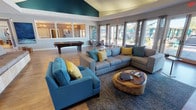 This well-appointed single-floor community space is designed for social gatherings and relaxation, combining modern aesthetics with functional amenities. The main lounge area is anchored by soaring vaulted ceilings that rise dramatically above a bold teal accent wall, creating a striking visual focal point. Large-format abstract artwork adorns the walls, complementing the contemporary color palette of deep teal, warm gray, and mustard yellow throughout the space.

The seating area is generously furnished with plush sectional and accent sofas arranged around a live-edge wood coffee table, providing ample room for conversation and leisure. A wall-mounted flat-screen television with a stylish media console adds an entertainment element to the lounge. Warm hardwood-style flooring runs throughout, enhancing the inviting atmosphere.

An adjacent game room features a full-size billiards table beneath the same soaring ceiling line, framed by interior clerestory windows that allow natural light to filter between zones. A kitchenette area with counter seating, pendant lighting, and modern appliances supports casual dining and events.

Floor-to-ceiling glass doors and large windows line an entire wall, flooding the interior with natural light and offering direct views and access to a landscaped outdoor pool deck. Sheer drapery panels frame the expansive glass, adding elegance while softening the transition between indoors and outdoors.