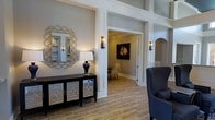 This refined single-floor space showcases a thoughtfully curated transitional design aesthetic that blends classic architectural details with contemporary elegance. The entryway sets a welcoming tone with warm wood-look tile flooring that flows seamlessly throughout, complemented by neutral gray walls that create a sophisticated yet inviting atmosphere.

Architectural highlights include crisp white millwork, decorative columns with transom detailing, and graceful arched doorways that add character and visual interest. Wall sconces provide warm, ambient lighting that enhances the upscale feel of the space.

The foyer is styled with a dark console cabinet featuring ornate mirrored panel doors, topped with a striking navy and white table lamp. A sculptural gold sunburst mirror adorns the wall, adding a glamorous focal point. A navy blue wingback accent chair with nailhead trim contributes a bold yet tasteful design element.

Beyond the entryway, an adjacent room draws the eye with a dramatic equestrian portrait, an elegant chandelier, and additional seating, hinting at the cohesive, gallery-like quality carried throughout. The overall ambiance is one of polished sophistication, combining rich textures, curated art, and well-considered details to create a space that is both functional and visually compelling.