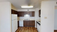 This single-floor residential unit offers a practical and comfortable layout designed for everyday living. The kitchen is well-equipped with dark wood cabinetry providing ample storage, light-toned countertops, and wood-look flooring. White appliances include a refrigerator, gas range, and dishwasher, complemented by a double-basin sink centered beneath under-cabinet lighting and a textured ceiling overhead. The living area features a ceiling fan with light fixture, wood-look flooring that transitions to plush, dark brown carpet, and a front entry door that allows natural light to enter the space. The bedrooms are generously sized, each finished with the same warm brown carpet and neutral walls, with windows fitted with blinds that allow for natural light and privacy. The bathroom includes a vanity with a mirrored medicine cabinet, dark wood cabinetry below, and bright overhead lighting. A full tub and shower combo with chrome fixtures and a safety grab bar completes the bathroom. Throughout the unit, textured ceilings and a consistent neutral color palette create a cohesive and understated aesthetic, offering a clean canvas well-suited for personalization.