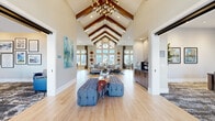 Located at 900 Elm Grove Ave in Gainesville, Georgia, this single-story community clubhouse offers a thoughtfully designed gathering space that blends modern elegance with inviting warmth. The centerpiece of the facility is a grand open lounge area distinguished by dramatic vaulted ceilings with striking exposed wood beams arranged in a chevron pattern and anchored by a bold geometric chandelier. Light wood-look flooring runs throughout, complementing the neutral palette and creating a cohesive, airy atmosphere. Large windows along the perimeter flood the space with abundant natural light and provide views of the surrounding community. Tufted upholstered benches and curated artwork add refined detail to the main lounge. A dedicated dining area features a substantial wood table with woven rope chairs, pendant lighting, and an elegantly tiled wet bar backdrop, ideal for social gatherings. A separate lounge zone includes a billiards table, wall-mounted television, and comfortable seating, offering a relaxed entertainment setting. The co-working area provides a marble-topped communal island, individual workstations, and pendant lighting, making it a functional space for productivity. Throughout the clubhouse, the design balances contemporary finishes with warm, textural elements, creating an environment that is both sophisticated and welcoming.