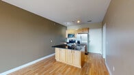 Located at 1500 River Shore Dr in Louisville, Kentucky, this single-floor residence offers a thoughtfully designed open-concept layout with a clean, contemporary aesthetic throughout. The kitchen serves as a functional centerpiece, featuring light maple cabinetry, dark granite countertops, a central island with an integrated sink, and a full suite of stainless steel appliances including a French door refrigerator, electric range, microwave, and oven. Track lighting illuminates the space, complementing the warm hardwood flooring that extends across the main living area. The open living space flows seamlessly from the kitchen and connects to a private balcony through a glass sliding door, offering views of the surrounding greenery and outdoor scenery. The bedroom is carpeted for comfort, features neutral warm-toned walls, a ceiling fan with light, and a window that allows natural light to filter in. A hallway with hardwood flooring connects the bedroom to the bathroom, which includes a wood-toned vanity with a white countertop and clean, simple fixtures. The bathroom also provides access to additional toilet and shower facilities. The overall color palette of warm taupes and greiges, paired with white trim, creates a cohesive and inviting atmosphere throughout the residence.