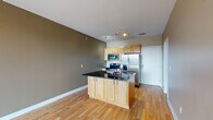 Located at 1500 River Shore Dr in Louisville, Kentucky, this single-floor residence offers a thoughtfully designed open-concept layout with a clean, contemporary aesthetic throughout. The kitchen serves as a functional centerpiece, featuring light maple cabinetry, dark granite countertops, a central island with an integrated sink, and a full suite of stainless steel appliances including a French door refrigerator, electric range, microwave, and oven. Track lighting illuminates the space, complementing the warm hardwood flooring that extends across the main living area. The open living space flows seamlessly from the kitchen and connects to a private balcony through a glass sliding door, offering views of the surrounding greenery and outdoor scenery. The bedroom is carpeted for comfort, features neutral warm-toned walls, a ceiling fan with light, and a window that allows natural light to filter in. A hallway with hardwood flooring connects the bedroom to the bathroom, which includes a wood-toned vanity with a white countertop and clean, simple fixtures. The bathroom also provides access to additional toilet and shower facilities. The overall color palette of warm taupes and greiges, paired with white trim, creates a cohesive and inviting atmosphere throughout the residence.
