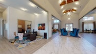 This single-story clubhouse is a well-appointed community space designed to impress through its architectural detail and thoughtful layout. The main entry and central corridor are defined by dramatic vaulted ceilings with exposed wood beam accents and a striking starburst chandelier that serves as a focal point. The flooring transitions between polished marble-look tile and herringbone-patterned wood-look planks, delineating functional zones throughout the space.

The reception and leasing area features dark wood desks set atop colorful geometric area rugs, with ample natural light filtering in through large windows with horizontal blinds. Adjacent to this is a comfortable lounge zone furnished with bold teal upholstered chairs and additional seating areas with plush orange sofas, teal rugs, and eclectic accent pieces.

A dedicated kitchenette area is outfitted with dark cabinetry, granite countertops, a sculptural geometric tile backsplash in deep blue, stainless steel appliances, and a long breakfast bar with stylish patterned counter stools illuminated by copper pendant lights. Additional private office or meeting rooms branch off the main corridor, offering versatile functionality within an overall warm and vibrant aesthetic.