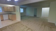This single-floor space offers an open and functional layout designed to maximize usability. The main living area is expansive, featuring wall-to-wall carpeting that extends throughout the space and provides a sense of warmth and continuity. Neutral, light-toned walls contribute to a clean and versatile atmosphere, ready to accommodate a variety of uses.

Adjacent to the main living area is a kitchen section, partially visible and equipped with wooden cabinetry and a countertop. A structural column and soffit detail define the boundary between the kitchen and living spaces, adding subtle architectural character without fully enclosing the areas.

A window positioned along one wall allows natural light to filter into the space, brightening the interior and offering a view of greenery outside. The ceiling is flat and features overhead lighting, ensuring the space remains well-lit.

Multiple doorways are visible throughout, leading to additional rooms or areas not fully visible in the current view, suggesting a thoughtful and connected floor plan. The overall ambiance is neutral and move-in ready, offering a blank canvas that is well-suited for customization to fit a range of needs and preferences.