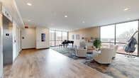 Located on the third floor of Alegria at 239 W King St in downtown Lancaster, PA, this elegantly appointed lobby lounge offers a welcoming and sophisticated communal environment. The space is defined by its open, airy layout with smooth, light-toned hardwood flooring that transitions into a plush, patterned area rug anchoring the main seating arrangement. A striking black grand piano serves as a focal centerpiece, positioned near floor-to-ceiling windows that frame sweeping views of the Lancaster cityscape, flooding the interior with abundant natural light. The seating area features a curated arrangement of woven armchairs and accent tables, creating an inviting atmosphere for conversation and relaxation. Warm beige walls are accented with vibrant artwork, adding a colorful, artistic character to the space. Recessed ceiling lighting complements the natural light throughout. On the opposite end of the lounge, two stainless steel elevator banks provide convenient access to the building, flanked by wall sconces and the prominently displayed Alegria 3 branding. The combination of modern finishes, thoughtful furnishings, a live-music-ready grand piano, and expansive urban views makes this third-floor lounge a distinguished and functional gathering space within the Alegria community.