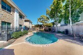 Located at 3333 Edenborn Ave in Metairie, Louisiana, this outdoor amenity area centers around a generously sized freeform swimming pool with clear, blue water. The pool features a curved, organic shape with entry steps and handrail access points on multiple sides, making it easily accessible. The surrounding concrete deck provides ample space for lounging, and is furnished with chaise lounge chairs and shaded seating areas with market umbrellas, ideal for relaxing under the Louisiana sun. A built-in spa or wading step feature is visible along one edge of the pool, adding an additional layer of comfort and leisure. The area is enclosed by a combination of wrought iron fencing and a tall wooden privacy fence, creating a secure and private retreat. Mature trees throughout the space offer natural shade and a lush, green backdrop against the clear blue sky. The pool area is flanked by multi-story brick and stone residential buildings, which lend a classic architectural character to the setting. Manicured landscaping with shrubs and ground cover lines the perimeter, enhancing the overall aesthetic. This well-appointed outdoor space offers a welcoming and functional environment for recreation and relaxation.