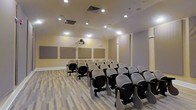 This single-floor space is designed as a dedicated lecture or presentation room, offering a functional and focused environment suited for educational sessions, seminars, or small conferences. The room features rows of theater-style seating with attached writing tablets, arranged to provide clear sightlines toward the front presentation area. A ceiling-mounted projector serves as the primary audio-visual tool, directed toward the front wall where multiple large acoustic panels are evenly spaced, aiding in sound management and reducing echo for improved audio clarity during presentations. The walls are finished in a warm neutral tone, contributing to a clean and professional ambiance. Recessed ceiling lighting provides uniform illumination throughout the space, supplemented by additional ceiling-mounted light fixtures that ensure consistent brightness. The flooring consists of light-toned wood-style planks, adding a contemporary touch to the room's overall aesthetic. The ceiling incorporates visible HVAC venting, suggesting a climate-controlled environment. The layout and furnishings collectively create a practical, purpose-built space optimized for focused learning and professional presentations.