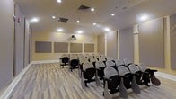 This single-floor space is designed as a dedicated lecture or presentation room, offering a functional and focused environment suited for educational sessions, seminars, or small conferences. The room features rows of theater-style seating with attached writing tablets, arranged to provide clear sightlines toward the front presentation area. A ceiling-mounted projector serves as the primary audio-visual tool, directed toward the front wall where multiple large acoustic panels are evenly spaced, aiding in sound management and reducing echo for improved audio clarity during presentations. The walls are finished in a warm neutral tone, contributing to a clean and professional ambiance. Recessed ceiling lighting provides uniform illumination throughout the space, supplemented by additional ceiling-mounted light fixtures that ensure consistent brightness. The flooring consists of light-toned wood-style planks, adding a contemporary touch to the room's overall aesthetic. The ceiling incorporates visible HVAC venting, suggesting a climate-controlled environment. The layout and furnishings collectively create a practical, purpose-built space optimized for focused learning and professional presentations.