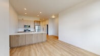 This single-floor unit showcases a clean, contemporary design defined by crisp white walls, light hardwood-style flooring, and an abundance of natural light. The open-concept layout seamlessly connects the kitchen and living area, creating a cohesive and functional living space. A hallway entry features a built-in niche, providing a practical landing spot upon arrival.

The kitchen is well-appointed with warm wood-toned cabinetry at both upper and lower levels, a large island with a white quartz countertop and undermount sink, and a full suite of stainless steel appliances including a gas range, microwave, dishwasher, and full-size refrigerator. Recessed lighting illuminates the kitchen and living area effectively.

The living area is generously proportioned with soaring double-height windows that flood the space with natural light and frame views of surrounding tree canopy. A sliding glass door opens directly to a private balcony, extending the living experience outdoors.

The bathroom maintains the unit's modern aesthetic with wood-toned cabinetry, a white quartz vanity top, a large mirror, and a glass-enclosed walk-in shower, all set against light wood-look tile flooring. A closet with sliding doors provides additional storage within the unit.