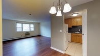 This 1-bedroom, 1-bathroom unit is thoughtfully laid out across a single floor, offering a comfortable and functional living environment. The open-concept design seamlessly connects the living area to the kitchen, creating a sense of spaciousness ideal for everyday living.

The living area features warm hardwood-style flooring, a large window that invites natural light and provides views of the surrounding neighborhood, and a wall-mounted air conditioning unit alongside baseboard heating for year-round comfort. An elegant three-light chandelier with bell-shaped glass shades serves as a decorative focal point, adding a refined touch to the space.

Transitioning into the kitchen, the flooring shifts to light-toned ceramic tile, providing a clean contrast. The kitchen is equipped with warm oak cabinetry offering generous storage, a white refrigerator, a dishwasher, and a sink positioned along the back wall with a dark tile backsplash. A flush-mount ceiling light ensures adequate task lighting throughout the kitchen area.

The neutral wall tones throughout the unit contribute to a bright, welcoming ambiance that is easy to personalize and furnish.