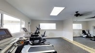 This fitness center offers a practical and well-maintained workout environment designed to support a range of cardio-focused exercise routines. The space is finished with neutral white walls and a durable rubber or carpet-style flooring, providing both comfort and functionality underfoot during workouts. Natural light filters in through horizontal windows along the walls, complementing a large recessed LED panel light in the ceiling that ensures consistent, bright illumination throughout the room. The equipment lineup includes treadmills, an elliptical cross-trainer, and a recumbent-style cardio machine, all arranged to allow comfortable spacing between users. The clean, minimalist interior design keeps the focus on functionality, with an uncluttered layout that makes navigation easy. A mirrored or glass panel along one wall adds a sense of openness to the space. The overall ambiance is bright, clean, and straightforward, making it a convenient and accessible option for residents or guests looking to maintain their fitness routine.