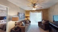 This single-floor residence features a comfortable living room designed with both functionality and warmth in mind. The space is anchored by plush wall-to-wall carpeting that adds a soft, inviting feel underfoot. A classic five-blade ceiling fan with an integrated light fixture serves as a central focal point overhead, providing both airflow and ambient illumination throughout the room.

The living area is furnished with a generously sized sofa adorned with decorative throw pillows, complemented by a matching armchair — both upholstered in rich brown tones that contribute to the room's warm, earthy color palette. Neutral beige walls are accented by warm ambient lighting from table lamps, creating a relaxed atmosphere.

A large sliding glass door dressed with vertical blinds and sheer curtain valances allows an abundance of natural light to flow into the space while offering a glimpse of a lush outdoor view. This feature enhances the sense of openness within the room. Decorative artwork on the wall adds a personalized touch, rounding out a living area that feels both practical and welcoming.
