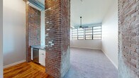 Located at 475 Harris Ave in Providence, Rhode Island, this single-floor unit showcases a compelling mix of industrial heritage and contemporary design. The open-concept living area is a standout, anchored by prominent exposed brick columns that serve as a visual centerpiece, contrasting handsomely with clean white walls and neutral-toned carpeting. A ceiling fan with a pendant-style fixture hangs above, while a bank of large, grid-paned windows floods the space with natural light and provides views of the surrounding neighborhood.

The kitchen is integrated seamlessly into the open layout, featuring white cabinetry, a dark countertop, and stainless steel appliances, including a dishwasher and refrigerator. Exposed ductwork along the ceiling reinforces the industrial aesthetic, while a wall-mounted mini-split unit provides climate control. A dedicated utility area includes a stacked washer and dryer, adding everyday convenience.

Additional rooms feature warm hardwood flooring, neutral gray walls, and angled ceiling details, contributing a sense of architectural interest. The consistent use of clean lines, updated fixtures, and original structural elements creates a cohesive, loft-inspired ambiance throughout the unit.