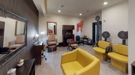 This full-service hair salon is thoughtfully designed to accommodate a wide range of beauty and grooming services within a single, efficiently organized space. The interior features a contemporary aesthetic with neutral walls, light tile flooring, and rich dark wood cabinetry and trim that create a warm, professional atmosphere.

The main styling area is anchored by bold yellow styling chairs positioned in front of a dedicated station with a framed mirror set against a textured, dark patterned accent wall. Product storage and tools are neatly arranged at each station. A dedicated shampoo area features a built-in wash unit with cabinet storage above, accompanied by a reclining shampoo chair and a gilded wall mirror, lending a classic touch to the space.

A pedicure station with a foot basin and reclining chair provides a comfortable setting for nail care services. The hooded dryer area offers a pair of yellow cushioned dryer chairs side by side, ideal for processing treatments in comfort. A small desk area with a computer provides a workspace for scheduling or client management. Vibrant abstract artwork in warm orange and red tones is displayed throughout, adding an artistic flair. Recessed lighting ensures the entire space is well-illuminated, enhancing the functional and welcoming environment.