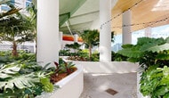 Located at 2200 NW 1st Ave in Miami's dynamic Wynwood neighborhood, this expansive outdoor amenity level is a masterfully designed open-air environment that seamlessly blends tropical landscaping with contemporary architecture. The space is organized into several distinct zones, each offering a unique experience. Sweeping curved planter beds, filled with lush tropical flora including monstera, palms, bromeliads, and banana-leaf plants, wind organically throughout the space, creating a garden-like atmosphere in the heart of the city. A covered lounge area features a bold, geometric orange canopy ceiling accented by string lights, with artificial turf flooring and casual bean bag seating — ideal for relaxed gatherings. Adjacent to this is an open-air seating terrace furnished with natural wood and linen chairs, sculptural accent pieces, and a striking warm-toned overhead canopy, all framed by panoramic views of the Miami skyline. A serene walkway corridor, lined with floor-to-ceiling gridded glass façades and trailing vines, connects the various zones with an airy, gallery-like quality. Throughout, white structural columns, pebble-inlaid tile pathways, and abundant natural light reinforce a clean, modern aesthetic that harmonizes with the vibrant tropical surroundings.