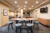 This single-floor community amenity space is thoughtfully designed to accommodate a variety of social and recreational activities. The layout flows seamlessly across several distinct zones, beginning with a generously sized dining area furnished with an assortment of seating options, including plaid-upholstered stools at long communal tables, cushioned armchairs, and a built-in banquette with warm leather-toned upholstery. Framed vintage-style artwork adds personality throughout the space, while recessed lighting and tray ceilings lend a polished, finished aesthetic. Natural light filters in through multiple windows fitted with horizontal blinds, offering views of surrounding greenery.

The full kitchen is equipped with stainless steel appliances — including a refrigerator and range with hood — dark cabinetry, and an expansive light-toned countertop island with bar seating, illuminated by a row of industrial-style pendant lights. A separate games alcove features rich hardwood-style flooring, a shuffleboard table as its centerpiece, and a trio of matching pendant lights overhead, creating a dedicated and inviting entertainment nook. A semi-private nook with a bistro table and stools offers a quieter seating option with an outdoor view. Double glass doors at one end open to an exterior patio area, further extending the social potential of this versatile space.