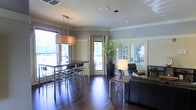 This single-floor space presents a thoughtfully designed open-concept layout that seamlessly combines a dining area and a lounge, creating a versatile and inviting environment. Rich, dark hardwood floors run throughout, adding warmth and a sense of sophistication to the interior. The dining area features a sleek, high bar-style table paired with modern metal stools, illuminated by a contemporary rectangular pendant light that anchors the space with an industrial-chic aesthetic. Large windows dressed with neutral linen curtains flood the room with natural light, enhancing the airy atmosphere. The lounge area is furnished with leather seating arranged around a low-profile coffee table, complemented by a stylish tripod floor lamp that adds a soft, ambient glow. A tall potted plant introduces a touch of greenery, bringing life and contrast to the neutral color palette of warm grays and whites. French-style glass panel doors provide an elegant transition, while recessed ceiling lighting ensures the space remains well-lit throughout. The overall ambiance is modern and refined, balancing comfort with clean, contemporary design elements that make this a functional and attractive multi-purpose space.
