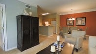 This single-story residence showcases an open-concept floor plan that seamlessly integrates the living, dining, and kitchen areas into one cohesive space. The living area is anchored by plush, neutral-toned carpeting and features a striking dark wood armoire with ornate carved medallion detailing, adding a traditional elegance to the room. A dark coffee table with candle accents completes the seating area.

Adjacent to the living space, the dining area is defined by a bold terracotta accent wall, which provides a warm, inviting contrast to the neutral tones throughout. A chandelier with candle-style lighting hangs above the dining table, which is surrounded by wicker-style chairs, creating a casual yet refined atmosphere.

The kitchen is positioned at the rear of the open layout, featuring wood-toned cabinetry, a peninsula countertop, and standard appliances including a refrigerator and stove. Overhead fluorescent lighting ensures a bright, functional workspace. Crown molding runs throughout the main living areas, adding architectural polish. The overall ambiance is warm and comfortable, with a cohesive blend of classic and casual design elements.