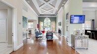 This sophisticated clubroom in Annapolis, Maryland offers a spacious, single-floor layout designed for both comfort and community. The space makes an immediate impression with its dramatic double-height ceilings adorned with an intricate coffered pattern, recessed lighting, and a statement pendant chandelier that anchors the main living area. Rich hardwood flooring flows throughout, complementing the soft sage green accent walls and crisp white millwork and baseboards. The main lounge area is generously proportioned, featuring multiple seating arrangements centered around a sleek fireplace with a wall-mounted flat-screen television above — perfect for relaxed gatherings. A striking arched window floods the space with natural light and frames lush outdoor greenery and views of a pool area beyond. The entry foyer transitions seamlessly from large-format tile flooring to hardwood, welcoming visitors with a chandelier, built-in window bench seating, frosted glass French doors, and vibrant wall art. Additional highlights include high clerestory windows that amplify the sense of volume, a secondary lounge nook with bistro-style high-top seating, a refreshment area, and wall-mounted displays. The overall ambiance is bright, polished, and inviting — a well-appointed community retreat suited for socializing or unwinding.