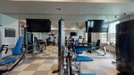 This single-level fitness center offers a comprehensive workout environment designed to accommodate a variety of exercise routines. The space is outfitted with a solid selection of cardio equipment, including treadmills, elliptical trainers, and stationary bikes, positioned to face wall-mounted flat-screen televisions that provide entertainment during workouts. Strength-training machines with blue upholstered seats and pads are distributed throughout, offering users options for targeted muscle conditioning.

Full-length mirrored walls line portions of the room, visually expanding the space and allowing users to monitor their form during exercise. The flooring features a distinctive checkered pattern in neutral tones, adding a clean, modern aesthetic. Recessed lighting keeps the space well-lit, complementing the natural light that filters in through large windows overlooking a landscaped outdoor area with palm trees and surrounding buildings.

A glass-paneled entry door provides access to the fitness center, and the layout is open and organized, ensuring ease of movement between equipment. The overall ambiance is bright, functional, and modern, creating an inviting environment well-suited for daily fitness use.