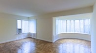 This single-floor space makes a strong first impression with its clean, open layout and timeless architectural details. The living area is anchored by richly patterned parquet hardwood flooring that adds warmth and character to the room, its geometric design catching the light beautifully throughout the day. Natural light is a defining feature of this space, pouring in generously through multiple large windows dressed with vertical blinds, creating a bright and airy atmosphere. A standout architectural element is the elegant curved bay window alcove, which adds a distinctive, classic charm to the room and creates an inviting nook that further enhances the sense of openness. The walls are finished in a soft, neutral tone that complements the warm flooring and contributes to the overall light and welcoming ambiance. Understated baseboard heating units line the walls beneath the windows, blending seamlessly into the design. The open floor plan offers flexibility in how the space can be arranged and utilized, making it well-suited for a variety of furniture configurations. Overall, the space combines classic architectural character with a bright, functional layout.