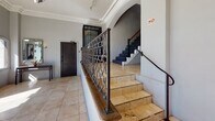 Located on Beverly Boulevard in Los Angeles, this distinctive building lobby showcases a compelling blend of classic architectural character and functional design. The main entry features a grand arched glass doorway that floods the space with abundant natural light, offering street-level views of the surrounding urban landscape. Large tile flooring runs throughout the ground level, lending a clean, polished foundation to the interior.

Intricate ornamental crown molding lines the upper perimeter of the lobby, a hallmark of the building's period-inspired aesthetic. A striking wrought iron railing with an elaborate scrollwork pattern borders the staircase landing, serving as a bold visual focal point. The staircase itself transitions from warm stone-tiled steps at the lower level to carpeted treads above, with a classic wood handrail guiding the ascent.

The upper landing continues the refined atmosphere, featuring an elegant chandelier, additional decorative crown molding, and arched architectural details that echo the ground floor's design language. Framed artwork and decorative furnishings add warmth to the neutral gray walls throughout. Practical elements such as a fire safety system and an elevator are also present, ensuring the space meets modern building standards while preserving its timeless character.
