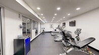 Located in Millcreek, Utah, this dedicated fitness center offers a thoroughly equipped exercise environment designed to support a wide range of workout routines. The space is laid out on a single level, providing a comfortable and functional footprint for both cardio and strength training activities.

The room features a clean, modern aesthetic with white walls, dark-trimmed doorways, and durable rubber flooring well-suited for high-traffic athletic use. Recessed ceiling lighting is distributed evenly throughout, ensuring the space is bright and well-illuminated at all times.

Cardio equipment includes multiple treadmills, a rowing machine, a stationary spin bike, and an elliptical trainer, catering to a variety of aerobic exercise preferences. For strength training, the facility is outfitted with a cable machine, a multi-position adjustable bench, a full rack of dumbbells, and TRX suspension straps mounted to the wall. A wall-mounted flat-screen television provides entertainment during workouts.

Additional amenities include a built-in water fountain with a bottle-filling station, a beverage refrigerator, and a conveniently located restroom. Large glass panels along one wall open sightlines into an adjacent area, adding a sense of openness to the environment. Overall, this fitness center is a practical and well-appointed facility suited for daily use.