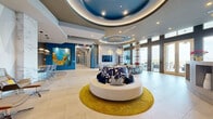 Located in Houston's prestigious Post Oak corridor, this sophisticated single-floor lobby space spans an impressively generous footprint, delivering a striking first impression through its thoughtful architectural design and vibrant interior palette. The space is defined by a series of dramatic tiered coffered ceilings, highlighted in deep blue lacquer and outfitted with recessed lighting and sculptural pendant chandeliers that cast a warm, inviting glow throughout. Large-format tile flooring transitions seamlessly across zones, complementing the open, airy layout. A standout feature is the floor-to-ceiling marble accent wall, which serves as a bold focal point near the elevator bank, while teal and cobalt blue structural columns punctuate the space with bursts of color. Curated large-scale abstract artwork adorns the walls, reinforcing a gallery-like atmosphere. The lounge area is anchored by a circular seating arrangement atop a gold area rug, surrounded by additional conversational seating groupings. A built-in media wall and bar/lounge area with green cabinetry offer functional amenities. A sleek wood-paneled reception desk provides a welcoming point of arrival. Floor-to-ceiling windows along the perimeter flood the interior with natural light and offer views of the surrounding Houston skyline, creating a seamless connection between the refined interior and the vibrant city beyond.