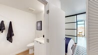 This single-floor unit in Berkeley, California presents a cohesive modern design with clean lines and a neutral palette throughout. Light wood-tone flooring runs continuously across all areas, contributing to a unified, airy aesthetic.

The kitchen is outfitted with floor-to-ceiling white cabinetry, open display shelving, and a full suite of stainless steel appliances including a refrigerator, electric range, microwave, and dishwasher. A single-basin sink and white quartz-style countertops complete the functional, minimalist layout.

The living area is bright and open, featuring a wall-mounted mini-split HVAC unit and a frosted glass panel door that adds a subtle architectural detail. The space flows naturally from the kitchen to a comfortable seating arrangement, creating a cohesive open-plan feel.

The bedroom is defined by distinctive shoji-style sliding panel doors with dark frames, lending a mid-century modern character. The bathroom is crisp and clean, featuring a white floating vanity with under-cabinet storage, a toilet, and a bathtub with wall-mounted hooks. Consistent white walls and wood-tone flooring maintain the design language found throughout the unit.

Overall, the space conveys a well-curated, contemporary sensibility with thoughtful storage solutions and cohesive finishes.
