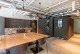 This single-floor commercial space showcases a thoughtfully designed interior that seamlessly merges industrial aesthetics with modern sophistication. Exposed ductwork, conduit pipes, and an open ceiling structure lend an industrial character throughout, while wide-plank light hardwood flooring grounds the space with warmth and texture.

The main area is anchored by a substantial solid-wood communal table surrounded by contemporary black chairs, making it ideal for collaborative work or meetings. Large windows along the perimeter flood the space with natural light and offer views of lush greenery outside. A dramatic navy accent wall with a built-in illuminated shelf and wall-mounted sconce lighting adds a bold visual focal point to the gathering area.

The kitchenette is designed with a dark, utilitarian palette, featuring stainless steel countertops, a dark tile backsplash, and matte charcoal cabinetry. It functions as a practical breakroom or prep station within the open-plan layout.

A set of full-height frameless glass double doors opens into a secondary room, offering visibility while maintaining a sense of enclosure — ideal for private meetings or focused work. Acoustic pod-style enclosures with textured cork or felt exteriors are also present, providing additional sound-isolated spaces. The overall ambiance is refined yet functional, suited for a contemporary professional environment.