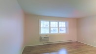 This single-floor space offers a clean, straightforward layout designed for comfortable living. The main room is finished with smooth hardwood flooring that reflects natural light, lending warmth and a polished appearance to the interior. A large, multi-pane double-hung window serves as a central architectural feature, flooding the room with ample daylight while offering views of a neighboring outdoor area, including what appears to be a deck or railing structure beyond. The walls are finished in a soft, neutral tone that enhances the bright and airy atmosphere of the space. Climate comfort is well-addressed, with a baseboard heating system running along the exterior wall beneath the window, complemented by a wall-mounted air conditioning unit for year-round temperature control. The overall design is modest and functional, with clean lines and minimal ornamentation, making it an adaptable space suitable for a variety of uses. The single-floor configuration ensures ease of accessibility and movement throughout.