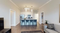 This single-floor residence showcases an open-concept layout that seamlessly connects the kitchen and living areas, creating a bright and inviting atmosphere. The kitchen is a standout feature, centered around a large island with a granite countertop and seating for four, making it ideal for casual dining and entertaining. White shaker-style cabinetry lines the walls, complemented by a subtle tile backsplash and stainless steel appliances, including a French door refrigerator with ice dispenser, a built-in microwave, and a range. Pendant lighting hangs above the island, adding a modern touch alongside a flush ceiling fixture. Crown molding runs throughout, lending a refined, polished look to the space. The flooring is warm-toned hardwood-style planks that extend throughout the open area, tying the kitchen and living spaces together cohesively. Neutral wall tones enhance the airy feel, while doorways on either side suggest access to additional private rooms. The overall ambiance is clean, contemporary, and functional, designed to balance everyday livability with stylish, modern finishes.