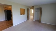 This single-floor space presents a clean, functional layout with neutral design elements throughout. The main room is finished with wall-to-wall carpet in a light beige tone, complemented by soft green-tinted walls and white baseboards, creating a calm and understated atmosphere. A notable architectural detail is the pass-through window cut into one wall, which connects to an adjacent room and adds a degree of openness to the layout. A built-in closet with bifold doors is positioned along one wall, providing convenient and organized storage with a hanging rod and shelf. A doorway leads to a secondary room visible in the background, which features a baseboard heater and access to natural light through a window, suggesting the space extends comfortably beyond the main room. The ceiling is smooth and standard in height, keeping the overall feel modest and practical. The neutral color palette and carpeted flooring throughout give the space a cohesive, move-in-ready appearance suited to a variety of uses. The layout is straightforward, maximizing usable floor space while incorporating essential storage and connectivity between rooms.