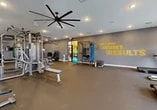 This single-floor fitness center is designed to support a full range of workout routines in a clean, modern environment. The space is finished with grey-painted walls, recessed lighting, and durable dark rubber flooring throughout, creating a functional and focused atmosphere. A large, industrial-style ceiling fan is prominently mounted overhead, ensuring consistent airflow across the workout floor.

The strength training area is well-appointed, featuring a multi-station cable machine, a Smith machine with a weight bench, a full rack of dumbbells displayed on tiered racks, and a functional training rig stocked with resistance bands, medicine balls, and stability balls. A large wall-mounted mirror alongside roller shades and multiple windows allows natural light to fill the space while providing visual feedback during workouts.

The cardio section lines one wall and includes treadmills, elliptical machines, stair climbers, and upright stationary bikes, each positioned in front of wall-mounted televisions for entertainment during exercise. A dedicated free-weight and stretching area offers open floor space with plyo boxes and additional functional training accessories.

Motivational typography in bold yellow lettering — reading "Don't Make Excuses, Make Results" — adorns the accent wall, reinforcing the energetic and goal-driven ambiance of the space. Glass-panel entry doors connect to an adjoining area, adding a sense of openness.