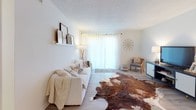 This single-floor residence offers a thoughtfully arranged layout encompassing multiple bedrooms, a living room, dining area, galley kitchen, and bathroom. The living room serves as a bright, welcoming centerpiece, featuring light hardwood-style flooring, a textured ceiling, and a large sliding glass door that fills the space with natural light. A plush white sofa, cowhide area rug, and decorative wall shelf create a cohesive, casual-modern aesthetic.

The dining area is distinguished by its earthy, bohemian character, with a round white table, wooden chairs, woven basket wall art, rustic floating shelves, and lush indoor plants adding warmth and texture. The galley kitchen is efficiently designed, featuring granite-style countertops, white cabinetry with brushed hardware, light hardwood-style flooring, and full appliances including a range and refrigerator.

The bedrooms each carry a calm, neutral palette with carpet flooring and blinds providing privacy and light control. One bedroom doubles as a creative workspace, accommodating a digital piano, desk, and recording equipment. The bathroom is neatly appointed with a granite-topped vanity, white cabinetry, and a tub-shower combination, maintaining the unit's consistent, understated aesthetic.