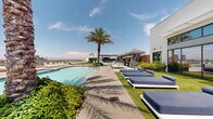 Located at 1010 E Orange St in Tempe, Arizona, this rooftop amenity deck delivers a resort-caliber outdoor experience in the heart of the city. The centerpiece is a generously sized swimming pool flanked by wide concrete pool surround and stretches of lush artificial turf, lined with rows of white-framed chaise lounges outfitted with navy blue cushions designed for extended relaxation under the Arizona sun. Towering palm trees punctuate the deck, providing natural shade and a tropical ambiance that complements the warm desert climate. A series of bold navy-and-white striped cabanas offer semi-private retreats, each equipped with outdoor seating and wall-mounted televisions for an elevated poolside experience. An open-air entertainment pavilion anchors one end of the deck, featuring a warm wood-paneled ceiling, built-in lounge sectionals, multiple mounted screens, and a ping pong table — ideal for social gatherings. The adjacent building façade is defined by expansive floor-to-ceiling glazing that reflects the pool and sky, reinforcing the seamless indoor-outdoor connection. Panoramic views of the Tempe and greater Phoenix skyline, along with the surrounding mountain ranges, provide a dramatic backdrop throughout the space.