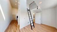 This efficiently designed studio in Seattle, WA offers a smart use of vertical space through a standout architectural feature: a soaring ceiling height in the living area paired with an elevated loft level accessed by a sleek black metal ladder with horizontal railing. Light wood flooring runs throughout the main level, lending warmth and continuity to the open layout. Natural light filters in through a window in the living area, brightening the clean white walls and reinforcing the airy feel of the space. The kitchenette is thoughtfully appointed with light wood cabinetry, white countertops, a stainless steel undermount sink, a two-burner induction cooktop, an over-the-range microwave, and a compact refrigerator — all the essentials neatly organized in a galley-style configuration. The bathroom features rich warm-toned hardwood flooring, a wood-front vanity with a sink, a full shower with a curtain rod, and a toilet, maintaining a consistent design language with the rest of the unit. Recessed lighting throughout provides consistent illumination, and the electrical panel is conveniently integrated into the main living space. The overall design is modern and minimal, making excellent use of every square foot.