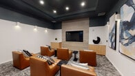 Located at 9840 Carroll Canyon Rd in San Diego, CA, this stylish screening room delivers a refined, theater-like atmosphere through thoughtful design and upscale furnishings. The space is defined by a striking dark tray ceiling that creates a dramatic, immersive ambiance, contrasted against crisp white lower walls adorned with elegant sconce lighting and a trio of large abstract art panels.

At the focal point of the room, a wide flat-panel display is mounted on a light wood accent wall, flanked by built-in low-profile cabinetry in a warm natural finish. Arranged in a theater-style layout, the seating consists of generously proportioned cognac leather lounge chairs paired with black marble side tables and an assortment of patterned throw pillows, offering both comfort and visual interest.

The floor is covered in a textured, patterned area rug that adds warmth and acoustic softness to the room. Recessed ceiling lights and cylindrical wall-mounted fixtures provide layered illumination throughout the space. Large glass-paneled doors connect the screening room to adjacent areas, offering glimpses of a broader communal environment. The overall design strikes a balance between cinematic functionality and contemporary lounge sophistication.