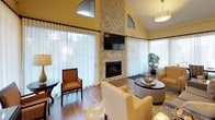 This single-story clubhouse is thoughtfully designed to provide a warm and inviting community gathering space. The centerpiece of the main lounge is a dramatic floor-to-ceiling stacked stone fireplace, topped with a wall-mounted flat-screen television, creating a striking focal point beneath soaring vaulted ceilings. Arched transom windows near the roofline allow additional natural light to fill the space, complementing the sheer floor-length curtains that line the surrounding windows and offer views of the lush greenery outside. Comfortable upholstered seating in warm golden tones is arranged to encourage conversation and relaxation, while hardwood-style flooring and soft yellow walls contribute to the overall inviting ambiance. Adjacent to the lounge is a fully equipped kitchen featuring rich dark cabinetry, stainless steel appliances including a range and dishwasher, light countertops, and open shelving for easy access. A dining table within the kitchen area provides a casual space for meals or gatherings. A separate activity area features ample table space bathed in natural light through expansive windows draped in sheer curtains, ideal for games, puzzles, or other leisure activities. Together, these spaces create a functional and comfortable clubhouse environment.