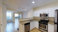 This single-floor residential space offers a well-designed, open-concept layout that seamlessly connects the kitchen to the living area, creating a functional and inviting atmosphere. The kitchen is a standout feature, outfitted with crisp white cabinetry, granite countertops, and a full suite of stainless steel appliances including a gas range, built-in microwave, and dishwasher. A double-basin sink is positioned along the counter, offering a practical workspace for everyday use. Track lighting runs along the kitchen ceiling, providing focused illumination throughout the space. Light-toned hardwood flooring extends throughout, contributing to a clean and cohesive aesthetic. The open sightline from the kitchen into the adjacent living area enhances the sense of spaciousness. Large sliding glass doors at the far end of the living area flood the interior with natural light and offer direct views of lush outdoor greenery, creating a pleasant indoor-outdoor connection. The overall ambiance is bright, modern, and functional, with neutral tones and contemporary finishes that give the space a versatile and well-maintained character.