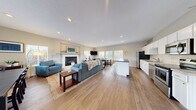 This single-floor space is designed around an expansive open-concept layout that seamlessly connects the living, dining, and kitchen areas, creating a cohesive and functional environment. Warm hardwood-style flooring runs throughout, lending continuity and a welcoming tone to the entire space.

The living area serves as the focal point, anchored by a well-appointed fireplace with a crisp white surround and decorative mantel. Recessed lighting illuminates the room, complemented by abundant natural light streaming through multiple windows fitted with white blinds. Vaulted ceilings above the fireplace wall add an airy sense of volume.

The kitchen is fully equipped with stainless steel appliances, including a refrigerator, range, microwave, and dishwasher. White shaker-style cabinetry pairs with light granite-style countertops, while a contrasting dark-topped island provides additional prep space and bar seating.

The dining area is positioned adjacent to the living space, featuring a large table and a bank of windows overlooking the outdoors, filling the area with natural light and outdoor views.

A separate lounge area, distinguished by vaulted ceilings and a bold striped area rug, offers flexible use with open shelving serving as a stylish room divider. The overall ambiance is clean, bright, and welcoming throughout.
