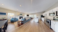 This single-floor space is designed around an expansive open-concept layout that seamlessly connects the living, dining, and kitchen areas, creating a cohesive and functional environment. Warm hardwood-style flooring runs throughout, lending continuity and a welcoming tone to the entire space.

The living area serves as the focal point, anchored by a well-appointed fireplace with a crisp white surround and decorative mantel. Recessed lighting illuminates the room, complemented by abundant natural light streaming through multiple windows fitted with white blinds. Vaulted ceilings above the fireplace wall add an airy sense of volume.

The kitchen is fully equipped with stainless steel appliances, including a refrigerator, range, microwave, and dishwasher. White shaker-style cabinetry pairs with light granite-style countertops, while a contrasting dark-topped island provides additional prep space and bar seating.

The dining area is positioned adjacent to the living space, featuring a large table and a bank of windows overlooking the outdoors, filling the area with natural light and outdoor views.

A separate lounge area, distinguished by vaulted ceilings and a bold striped area rug, offers flexible use with open shelving serving as a stylish room divider. The overall ambiance is clean, bright, and welcoming throughout.