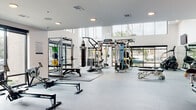 This contemporary fitness center is thoughtfully designed to accommodate a wide range of workout styles and fitness goals. The space is organized into distinct functional zones, each tailored to a specific type of training. The entry area sets an energetic tone with warm light wood flooring, bold abstract artwork, and sleek glass doors that invite natural light into the space.

The strength training zone is a standout feature, boasting soaring double-height ceilings, expansive floor-to-ceiling windows, and pendant lighting that creates an open, airy atmosphere. This area is outfitted with a multi-functional cable machine, a power rack with weight plates, a heavy bag for boxing training, adjustable benches, and a full dumbbell rack reflected in a large wall mirror. Wall-mounted flat-screen televisions are positioned for easy viewing throughout.

The cardio zone is equally well-appointed, lined with treadmills, ellipticals, air bikes, a StairMaster step mill, and a VersaClimber, all positioned to take advantage of the natural light streaming through large perimeter windows. Durable rubber and speckled flooring throughout provides safety and comfort underfoot.

A dedicated support area includes a water station and open cubbies for storage, finished with distinctive patterned accent wall tiles that add a playful, modern design element to the overall aesthetic.