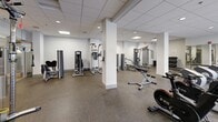 Located at 3333 Wisconsin Ave NW in Washington, DC, this fitness center offers a comprehensive, single-floor layout designed to support a full range of workout routines. The space is organized into distinct functional zones, making it easy to move between cardio training, strength work, and flexibility exercises.

The cardio section is lined with a row of elliptical trainers, treadmills, and stationary bikes, each equipped with individual monitors for entertainment or workout tracking. Adjacent to this area, a dedicated strength training zone houses multiple cable and weight machines, providing options for both isolated and compound movements. A tiered dumbbell rack with flat benches rounds out the free weights area.

A separate stretching and functional training zone features exercise mats, stability balls, and resistance equipment, complemented by a wall-mounted flat-screen TV. A colorful, motivational word-art mural adds energy and visual interest to this section of the room.

Throughout the space, durable rubberized flooring provides safety and comfort underfoot, while a drop ceiling with recessed panel lighting ensures consistent, even illumination. Full-length mirrors along one wall visually expand the room and assist with form checks during workouts. A water dispenser is conveniently accessible, and the overall layout allows for easy circulation between equipment stations. The clean, white walls and organized arrangement of equipment create a professional and focused fitness environment.