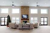This generously sized single-floor space is designed as an open, multi-use common area that balances comfort with functionality. The centerpiece of the main lounge is a dramatic floor-to-ceiling stacked stone fireplace fitted with a mounted flat-screen television and a rustic wood mantel, creating a warm and inviting focal point. Surrounding the fireplace, neutral-toned upholstered seating is arranged around a farmhouse-style coffee table, encouraging relaxed social interaction.

The soaring ceilings, lined with a row of clerestory windows along multiple walls, allow an abundance of natural light to fill the interior throughout the day. Dual ceiling fans circulate air efficiently across the expansive open floor plan, which is finished with neutral patterned carpet throughout. Exposed ductwork along the upper walls adds an industrial accent that contrasts with the otherwise warm, contemporary aesthetic.

Multiple exterior entry points, including glass-paneled double doors, connect the space to the outdoors. A quieter activity alcove features light wood tables and chairs, well-suited for games, puzzles, or small group gatherings. Additional open areas near entry corridors provide flexible space for various uses. The overall ambiance is bright, welcoming, and thoughtfully designed for communal living.